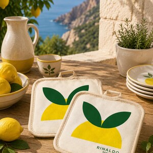 Puede incluir: Dos agarradores de tela de color blanco roto con un diseño gráfico de un limón amarillo y dos hojas verdes. El agarrador delantero muestra el texto RINALDO DESIGN en letras negras. Cada pieza incluye una pequeña trabilla de tela.