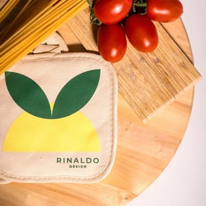 May include: A yellow and green lemon-shaped pot holder with the text "RINALDO DESIGN" on a wooden surface with a bunch of red tomatoes and a bundle of spaghetti.