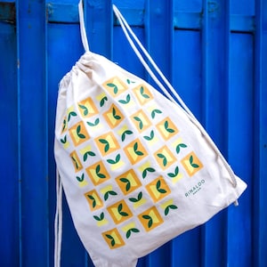 Könnte beinhalten: Eine weiße Kordeltasche mit einem gelben und grünen geometrischen Muster. Die Tasche hat den Text "RINALDO DESIGN" in der unteren rechten Ecke aufgedruckt.