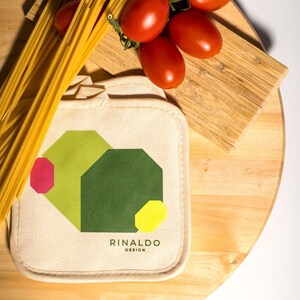 Op de afbeelding: Twee beige katoenen ovenwanten met een geometrisch ontwerp met groene, gele en rode vormen. De ovenwanten hebben de tekst "RINALDO DESIGN" erop gedrukt. De ovenwanten liggen op een houten oppervlak met spaghetti en tomaten.