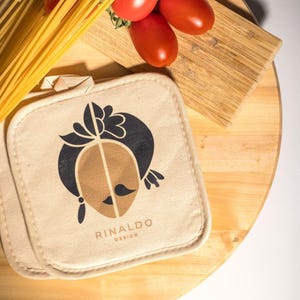 Könnte beinhalten: Ein beiger Topflappen mit einer braunen und schwarzen Grafik eines Gesichts mit einem Schnurrbart und einem floralen Design. Der Text "RINALDO DESIGN" ist unter der Grafik gedruckt. Der Topflappen liegt auf einer Holzoberfläche mit einem Bund roter Tomaten und einem Bündel Spaghetti.