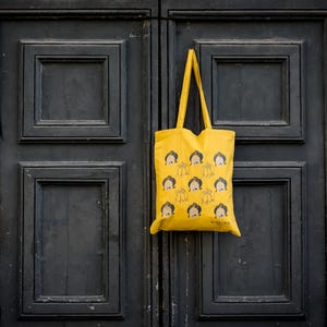 May include: A yellow tote bag with a repeating pattern of cartoon faces. The bag is hanging on a black wooden door.