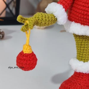 Puede incluir: Primer plano de una figura de ganchillo hecha a mano, roja con adornos blancos y extremidades verdes. La figura sostiene un adorno rojo y amarillo. El texto "olya_and_toys" es visible. Probablemente sea una decoraci&oacute;n navide&ntilde;a o festiva.