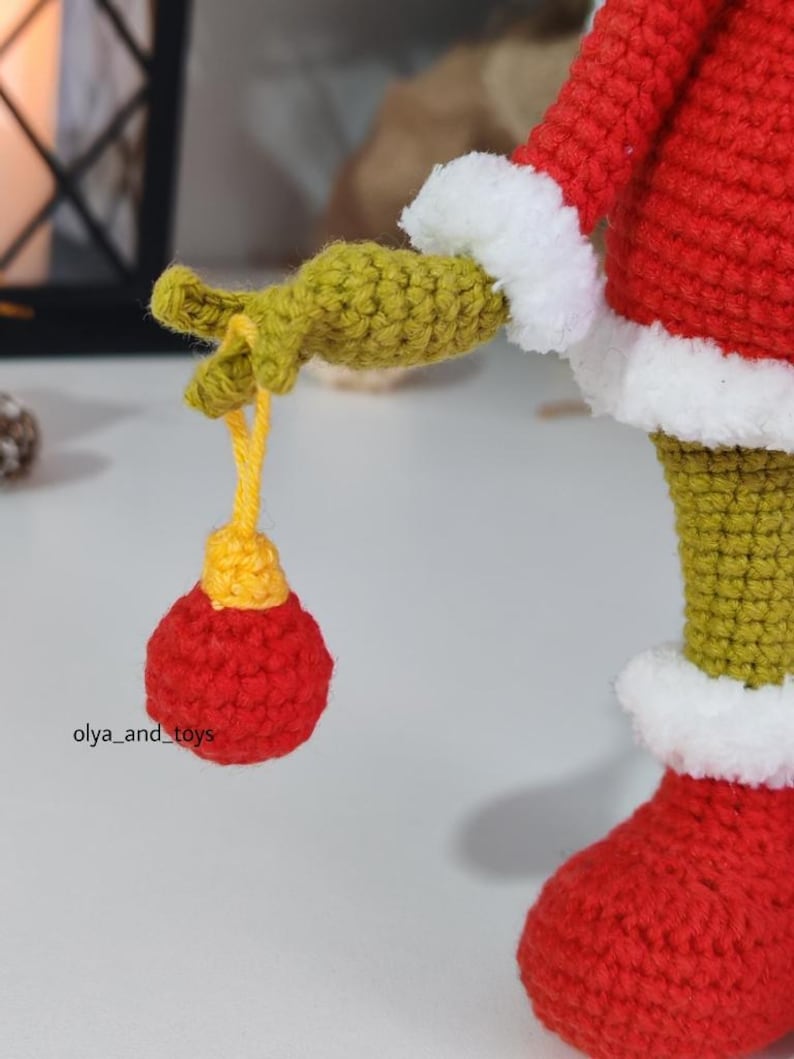 Puede incluir: Primer plano de una figura de ganchillo hecha a mano, roja con adornos blancos y extremidades verdes. La figura sostiene un adorno rojo y amarillo. El texto "olya_and_toys" es visible. Probablemente sea una decoraci&oacute;n navide&ntilde;a o festiva.
