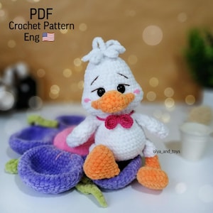 May include: A white crocheted duck with orange feet and beak, wearing a pink bow tie. The duck is sitting on a purple and pink crocheted flower.