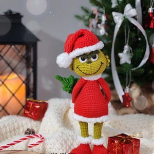 Puede incluir: Mu&ntilde;eco Grinch tejido a ganchillo con un traje y gorro de Pap&aacute; Noel rojos con ribetes blancos. El Grinch tiene una cara amarilla, ojos verdes y una sonrisa traviesa. Sostiene un peque&ntilde;o &aacute;rbol verde. El mu&ntilde;eco est&aacute; sobre una superficie blanca con adornos navide&ntilde;os.