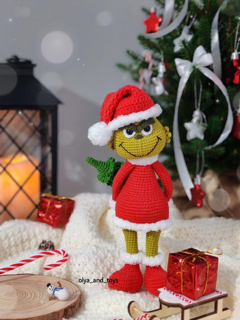 Puede incluir: Mu&ntilde;eco Grinch tejido a ganchillo con un traje y gorro de Pap&aacute; Noel rojos con ribetes blancos. El Grinch tiene una cara amarilla, ojos verdes y una sonrisa traviesa. Sostiene un peque&ntilde;o &aacute;rbol verde. El mu&ntilde;eco est&aacute; sobre una superficie blanca con adornos navide&ntilde;os.