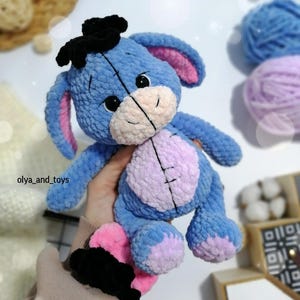 May include: A blue and purple crocheted stuffed animal resembling a donkey with a black bow on its head. The toy has pink ears and a stitched smile.