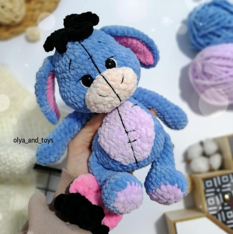 May include: A blue and purple crocheted stuffed animal resembling a donkey with a black bow on its head. The toy has pink ears and a stitched smile.
