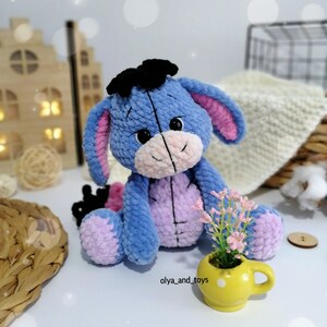 May include: A blue and pink crocheted stuffed animal resembling a donkey with black ears and a black bow. The toy is sitting on a white surface with a small yellow cup and pink flowers.