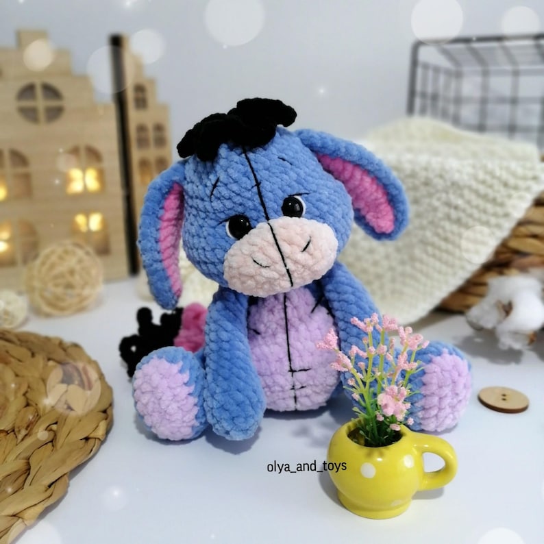 May include: A blue and pink crocheted stuffed animal resembling a donkey with black ears and a black bow. The toy is sitting on a white surface with a small yellow cup and pink flowers.