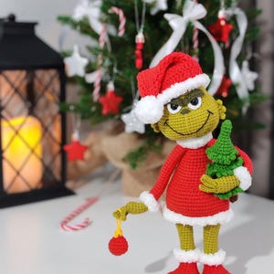 Puede incluir: Mu&ntilde;eco Grinch tejido a ganchillo con un traje de Pap&aacute; Noel rojo con ribetes blancos y un gorro a juego. El Grinch sostiene un peque&ntilde;o &aacute;rbol de Navidad verde y una bola roja. El mu&ntilde;eco tiene piel verde y una expresi&oacute;n traviesa.