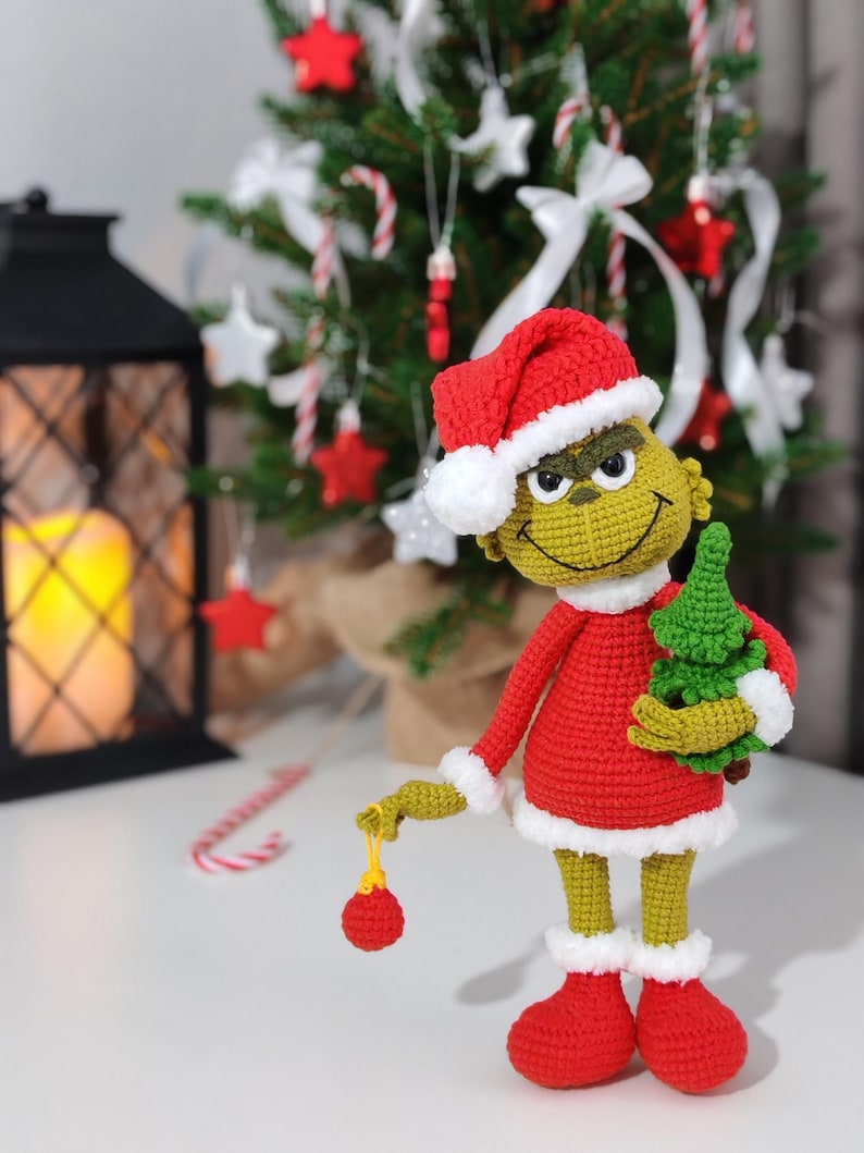 Puede incluir: Mu&ntilde;eco Grinch tejido a ganchillo con un traje de Pap&aacute; Noel rojo con ribetes blancos y un gorro a juego. El Grinch sostiene un peque&ntilde;o &aacute;rbol de Navidad verde y una bola roja. El mu&ntilde;eco tiene piel verde y una expresi&oacute;n traviesa.