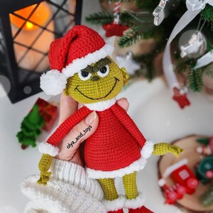 Puede incluir: Mu&ntilde;eco Grinch tejido a ganchillo con traje y gorro de Pap&aacute; Noel rojos con ribetes blancos. El Grinch tiene piel verde, una cara sonriente y sostiene un objeto tem&aacute;tico navide&ntilde;o. El mu&ntilde;eco mide unos 20 cm de alto.