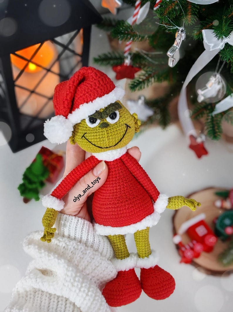 Puede incluir: Mu&ntilde;eco Grinch tejido a ganchillo con traje y gorro de Pap&aacute; Noel rojos con ribetes blancos. El Grinch tiene piel verde, una cara sonriente y sostiene un objeto tem&aacute;tico navide&ntilde;o. El mu&ntilde;eco mide unos 20 cm de alto.
