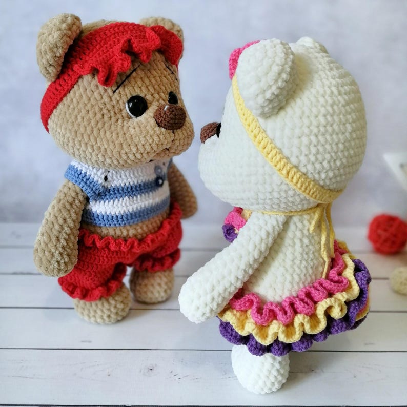 May include: Two crocheted teddy bears, one brown with a red headband and blue and white striped shirt, the other white with a yellow headband and a pink, yellow, and purple ruffled skirt.