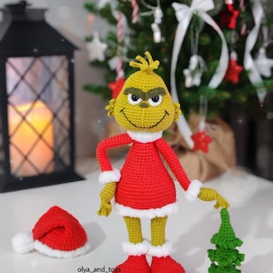 Puede incluir: Mu&ntilde;eco Grinch tejido a ganchillo con un atuendo rojo con ribetes blancos, sosteniendo un peque&ntilde;o &aacute;rbol de Navidad verde. Un gorro de Pap&aacute; Noel rojo con un pomp&oacute;n blanco est&aacute; cerca. El fondo incluye un &aacute;rbol de Navidad y una linterna decorativa.