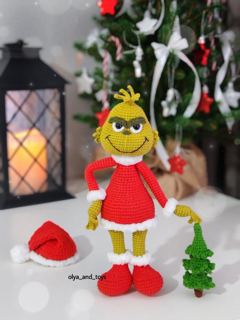 Puede incluir: Mu&ntilde;eco Grinch tejido a ganchillo con un atuendo rojo con ribetes blancos, sosteniendo un peque&ntilde;o &aacute;rbol de Navidad verde. Un gorro de Pap&aacute; Noel rojo con un pomp&oacute;n blanco est&aacute; cerca. El fondo incluye un &aacute;rbol de Navidad y una linterna decorativa.