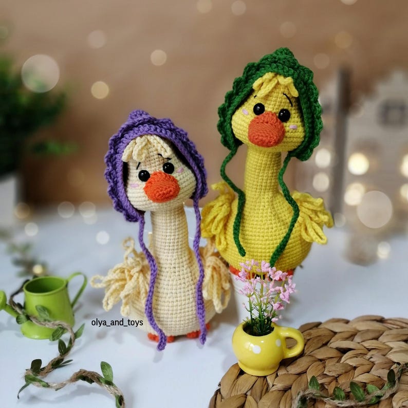 May include: Two crocheted yellow duck plushies with purple and green hats. The ducks are sitting on a woven surface with a small potted plant in front of them.