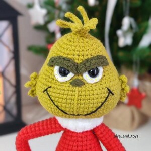 Puede incluir: Mu&ntilde;eca de ganchillo hecha a mano de un personaje verde con una expresi&oacute;n traviesa. La mu&ntilde;eca tiene ojos negros, una amplia sonrisa y un atuendo rojo. La cabeza de la mu&ntilde;eca es amarilla con cejas verdes y un mech&oacute;n de pelo.