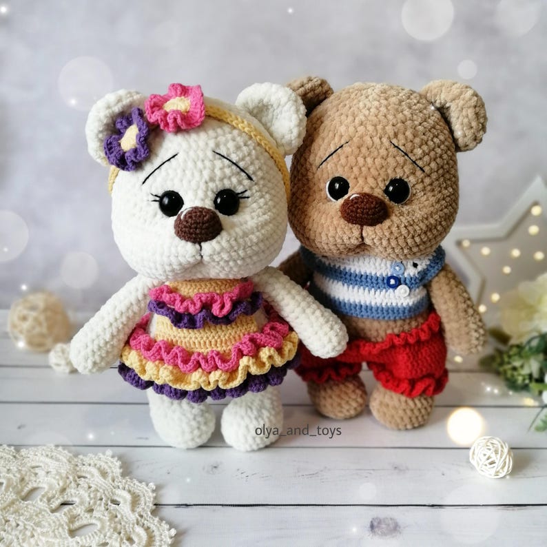 May include: Two crocheted teddy bears, one white with a yellow, pink, and purple dress and a flower headband, and one brown with a blue and white striped shirt and red pants.