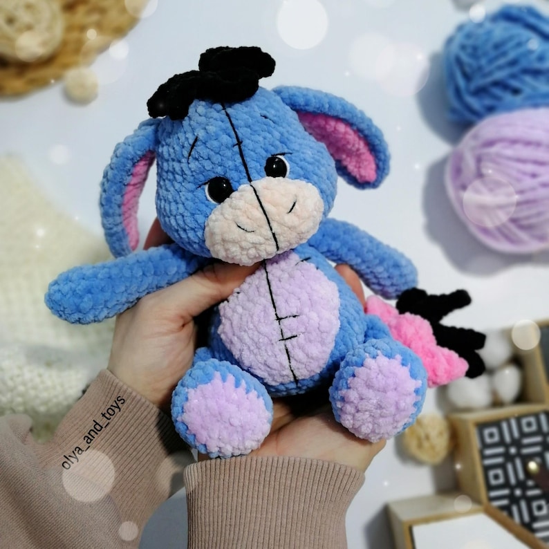 May include: A blue and pink crocheted stuffed animal donkey with black ears and a black bow. The donkey has a stitched smile and large black eyes.