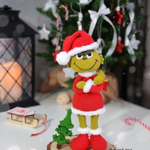 Puede incluir: Figura del Grinch de ganchillo con gorro de Pap&aacute; Noel, vestido y botas rojas con ribetes blancos. La figura tiene la piel amarilla y est&aacute; sobre una rodaja de madera con un peque&ntilde;o &aacute;rbol verde. Un &aacute;rbol de Navidad est&aacute; al fondo.