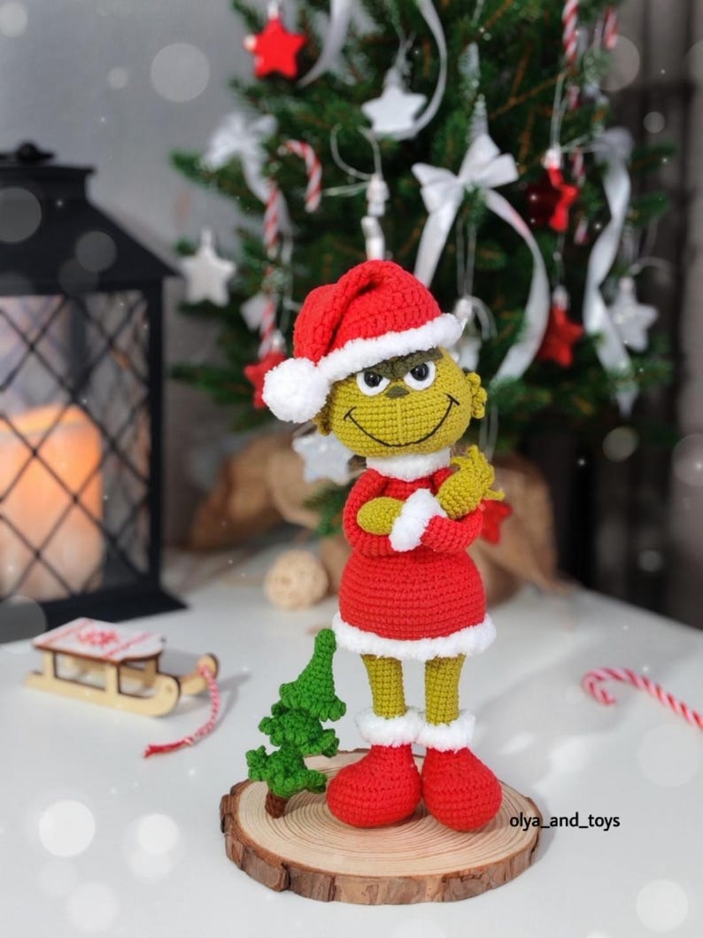 Puede incluir: Figura del Grinch de ganchillo con gorro de Pap&aacute; Noel, vestido y botas rojas con ribetes blancos. La figura tiene la piel amarilla y est&aacute; sobre una rodaja de madera con un peque&ntilde;o &aacute;rbol verde. Un &aacute;rbol de Navidad est&aacute; al fondo.