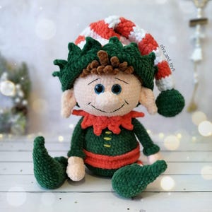 May include: A crocheted elf doll, featuring a green body, a red and white striped hat, and a red collar. The elf has a smiling face with blue eyes. This handmade doll is a festive holiday decoration.
