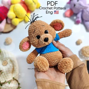 May include: A handmade brown crochet bear with a blue shirt and pink inner ears. The bear has black eyes and a black nose. The image includes the text "PDF Crochet Pattern Eng".