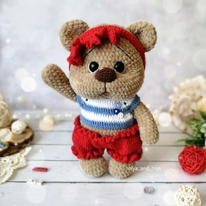 May include: A crocheted teddy bear wearing a red headband, a blue and white striped shirt, and red shorts. The bear has brown fur and large black eyes.