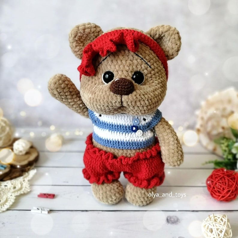 May include: A crocheted teddy bear wearing a red headband, a blue and white striped shirt, and red shorts. The bear has brown fur and large black eyes.