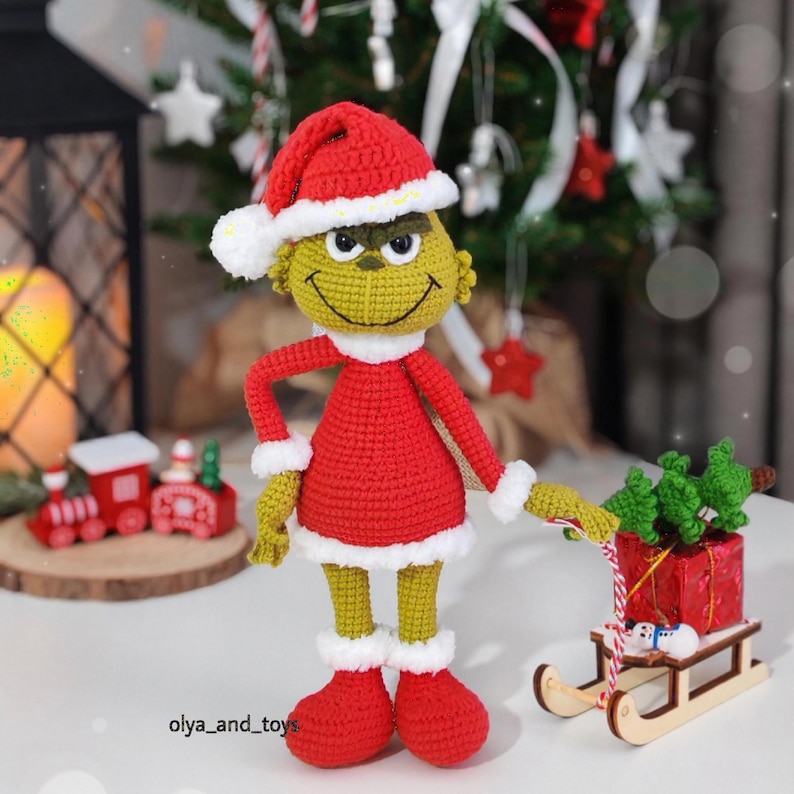 Puede incluir: Mu&ntilde;eco Grinch tejido a ganchillo con gorro y vestido rojos con ribete blanco. El Grinch tiene piel verde, una cara sonriente y botas rojas. Un peque&ntilde;o trineo de madera con un regalo y &aacute;rboles de Navidad est&aacute; junto al mu&ntilde;eco. El texto "olya_and_toys" es visible.