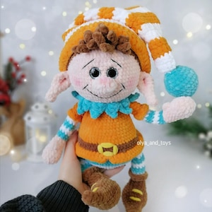 May include: A handmade crochet elf doll with a cheerful expression. The doll features an orange and white striped hat, orange tunic, blue collar, and brown boots. The doll is holding a blue ball and has the text "olya_and_toys".