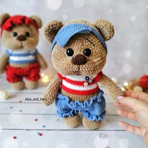 May include: Two crocheted teddy bears wearing sailor-themed outfits. The bear on the left is wearing a red and white striped shirt, red shorts, and a red headband. The bear on the right is wearing a red and white striped shirt, blue shorts, and a blue headband. Both bears have brown fur and black eyes.