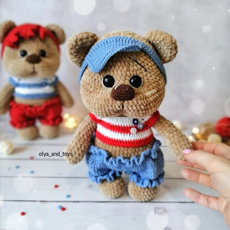 May include: Two crocheted teddy bears wearing sailor-themed outfits. The bear on the left is wearing a red and white striped shirt, red shorts, and a red headband. The bear on the right is wearing a red and white striped shirt, blue shorts, and a blue headband. Both bears have brown fur and black eyes.