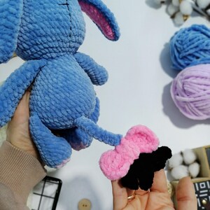 May include: A blue crocheted stuffed animal shaped like a bunny with a pink bow on its tail. The bunny is being held by a hand.