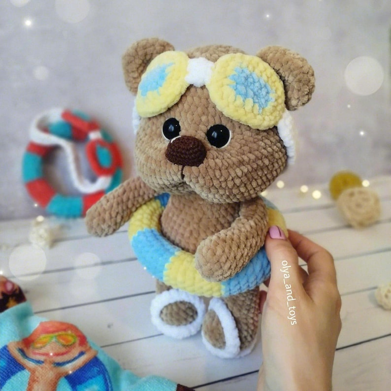 May include: A brown crocheted teddy bear wearing yellow and blue goggles and a blue and yellow life preserver. The bear is standing on a wooden surface.