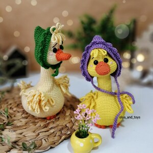 May include: Two crocheted yellow duck plushies wearing hats. One duck is wearing a green hat and the other is wearing a purple hat. The ducks are sitting on a woven basket.