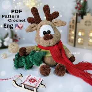 May include: A handmade crochet reindeer with brown antlers, a red scarf, and a green shirt. The reindeer is holding a green crochet Christmas tree. The image includes the text "PDF Pattern Crochet Eng".