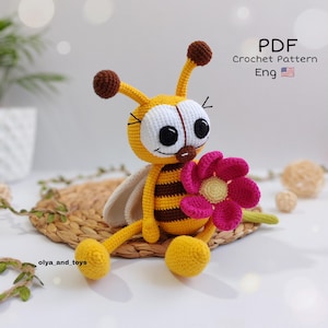 May include: A crocheted bee doll with a yellow body, brown stripes, and large white eyes. The bee holds a pink flower and has brown antennae. The image includes the text "PDF Crochet Pattern Eng".