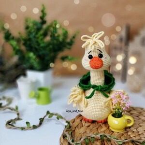 May include: A crocheted goose toy with a green hooded scarf, yellow beak, and black eyes. The goose is sitting on a woven basket with a small yellow flower pot next to it.
