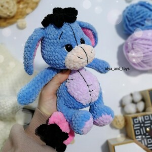May include: A blue crocheted stuffed animal with pink ears and a black bow. The animal has a pink and purple belly and black eyes. The stuffed animal is sitting on a hand.