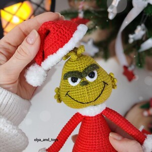 Puede incluir: Mu&ntilde;eco Grinch tejido a ganchillo, vestido con un traje rojo con ribetes blancos y un gorro de Pap&aacute; Noel a juego. El Grinch tiene una cara verde, cejas negras y una expresi&oacute;n sonriente. Decoraci&oacute;n navide&ntilde;a.
