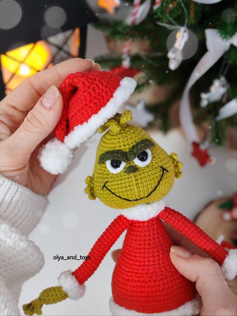 Puede incluir: Mu&ntilde;eco Grinch tejido a ganchillo, vestido con un traje rojo con ribetes blancos y un gorro de Pap&aacute; Noel a juego. El Grinch tiene una cara verde, cejas negras y una expresi&oacute;n sonriente. Decoraci&oacute;n navide&ntilde;a.