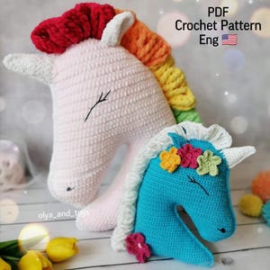 Könnte beinhalten: Zwei gehäkelte Amigurumi-Einhorn-Spielzeuge. Das größere Einhorn ist rosa mit einer Regenbogenmähne und einem weißen Horn. Das kleinere Einhorn ist blau mit einem weißen Horn und einer Blumenkrone mit gelben, grünen und rosa Blumen.