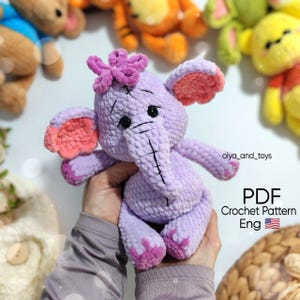 Könnte beinhalten: Ein handgefertigtes, lavendelfarbenes Häkelelefanten-Spielzeug mit rosa Akzenten an Ohren und Füßen und einer kleinen Blume auf dem Kopf. Das Spielzeug wird in den Händen einer Person gehalten. Der Text "PDF Crochet Pattern Eng" ist ebenfalls enthalten.