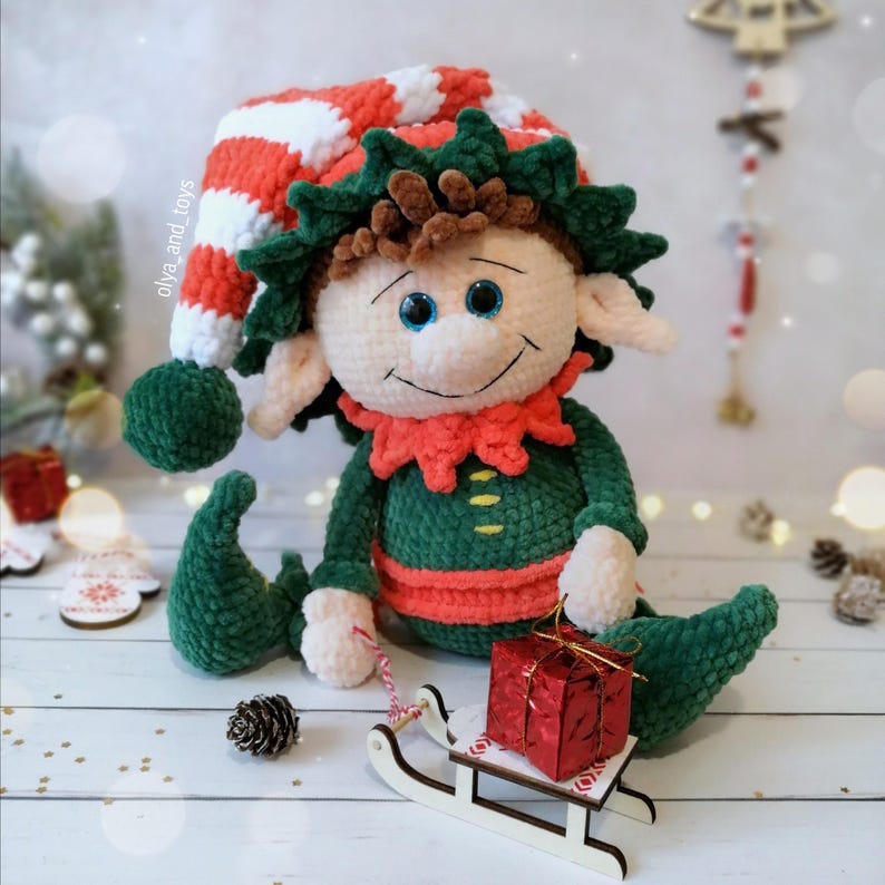 May include: A handcrafted Christmas elf doll wearing a red and white striped hat, green outfit, and a red collar. The elf is holding a small wooden sled with a red gift box. The doll has blue eyes and a smiling face.