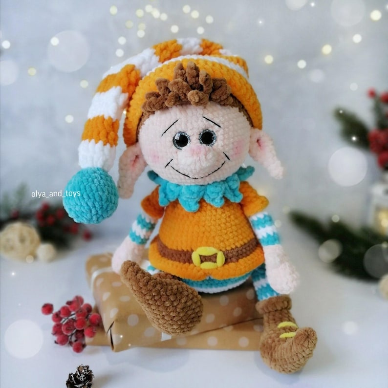May include: A handcrafted crochet elf doll with a smiling face. The elf is dressed in an orange and blue outfit, a striped hat, and brown boots. The doll is seated on a wrapped gift. The background is blurred with bokeh lights.