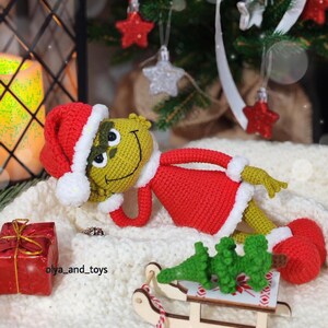 Puede incluir: Mu&ntilde;eco Grinch tejido a ganchillo, con gorro de Pap&aacute; Noel y traje rojo con ribetes blancos. Est&aacute; tumbado sobre una manta de punto blanca, con una peque&ntilde;a caja de regalo roja y un &aacute;rbol de Navidad en miniatura sobre un trineo. La cara es verde.